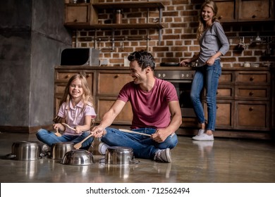 Cute Little Girl And Her Dad Are Using Wooden Spoons And Smiling While Playing Drums With Dishes In Kitchen, Mom Is Cooking