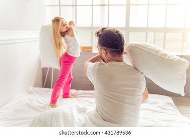Cute little girl and her father are fighting with pillows and smiling while sitting on bed at home