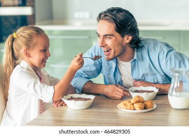 Cute little girl and her handsome father are smiling while having breakfast in kitchen at home. Girl is feeding her dad with a spoon