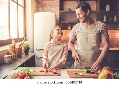 Cute little girl and her handsome dad are cutting vegetables and smiling while cooking in kitchen at home