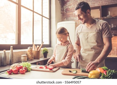 Cute Little Girl And Her Handsome Dad Are Cutting Vegetables And Smiling While Cooking In Kitchen At Home