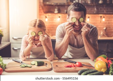 Cute Little Girl And Her Handsome Dad Are Holding Slices Of Cucumber And Smiling While Cooking In Kitchen At Home