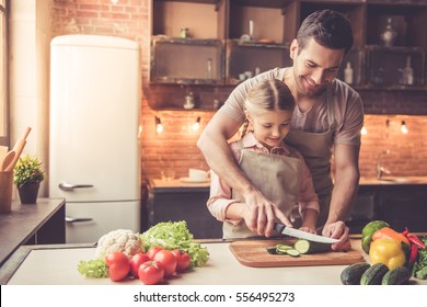 Cute Little Girl And Her Handsome Dad Are Cutting Vegetables And Smiling While Cooking In Kitchen At Home