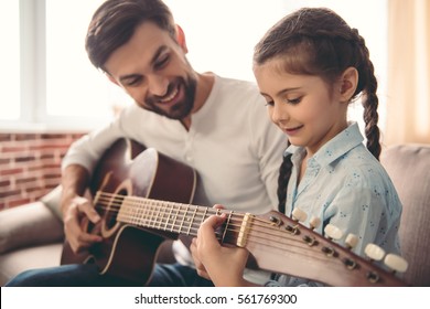 Cute little girl and her handsome father are playing guitar and smiling while sitting on couch at home