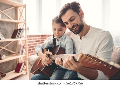 Cute little girl and her handsome father are playing guitar and smiling while sitting on couch at home - Powered by Shutterstock