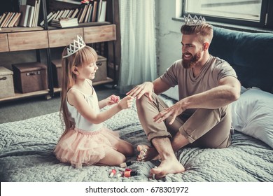 Cute Little Girl And Her Handsome Bearded Dad In Crowns Are Smiling While Playing In Her Room. Girl Is Painting Her Dad Nails