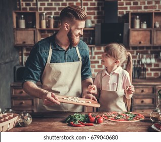 Cute Little Girl And Her Handsome Bearded Dad In Aprons Are Talking And Smiling While Cooking Pizza In Kitchen