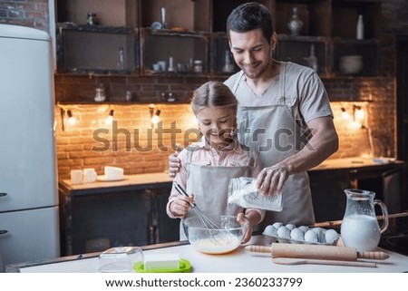 Cute little girl and her handsome dad in aprons are whisking eggs with flour and smiling while baking in kitchen at home