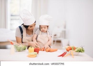 Cute Little Girl And Her Mom In Chef's Hats Are Cutting Vegetables Cooking A Salad And Smiling