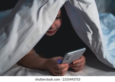 Cute Little Girl Hides Under Blanket With Smartphone At Night When Everyone Is Asleep. Child Is Smiling And Having Fun Watching Cartoons. Children. Blurred