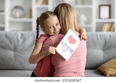 Cute Little Girl Holding Greeting Card With Drawn Red Heart And Embracing Her Mom At Home, Loving Female Child Greeting Mommy With Mother's Day While Bonding Together In Living Room Interior - Shutterstock ID 2311020347