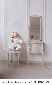 A Cute Little Girl Is Holding A Makeup Brush And Doing Make Up. Baby Girl Is Sitting On The Chair Near The Classic Mirror At . Kid's Fashion. Little Girl Fashionista.