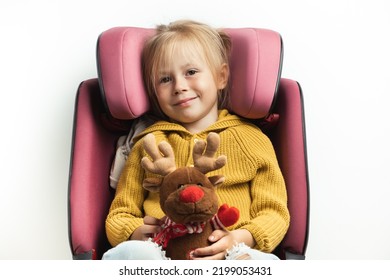 Cute little girl holding stuffed Rudolph Red-Nosed Reindeer toy while sitting in a safety car seat. Kid safety while traveling by car. Isolated on white. Merry Christmas. Holiday adventure concept