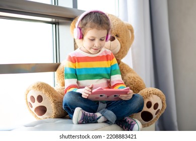 Cute little girl holding tablet computer in her hands. She is in headphones. Advertising new gadget, educational app. She is near the window. Games. Children use technology. - Shutterstock ID 2208883421