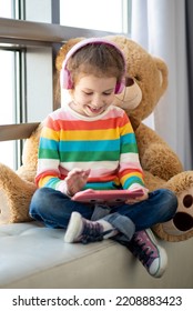 Cute little girl holding tablet computer in her hands. She is in headphones. Advertising new gadget, educational app. She is near the window. Games. Children use technology. - Shutterstock ID 2208883423