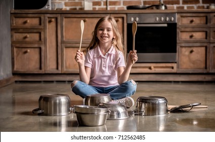Cute little girl is holding wooden spoons, looking at camera and smiling while playing drums with dishes in kitchen
