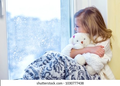 Cute Little Girl Hugging A Teddy Bear. A Cute Baby In The Room Sits At The Window In The Winter