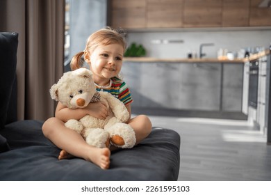 Cute little girl hugging teddy bear while sitting on sofa at home. High quality photo