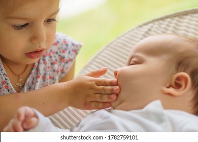 Cute little girl kissing his newborn sister. Toddler kid meeting new born sibling. Infant sleeping in white bouncer under a blanket. Kids playing and bonding. Children with small age difference.