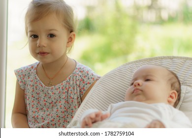 Cute little girl kissing his newborn sister. Toddler kid meeting new born sibling. Infant sleeping in white bouncer under a blanket. Kids playing and bonding. Children with small age difference.