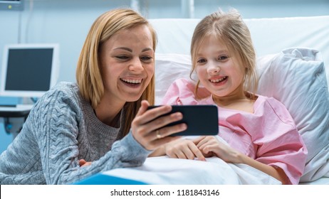 Cute Little Girl Lies on a Bed in the Children's Hospital, Her Mother Sits Beside, They Watch Cartoons/ Funny Videos on Smartphone. Modern Pediatric Ward. - Powered by Shutterstock