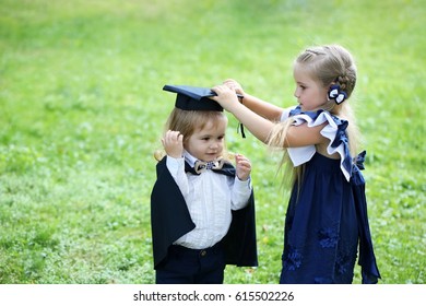 Cute little girl with long hair in blue dress dressing adorable small boy in black graduation hat or cap and robe on summer day outdoors on green grass background