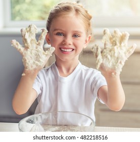 Cute little girl is looking at camera and smiling while kneading the dough in the kitchen