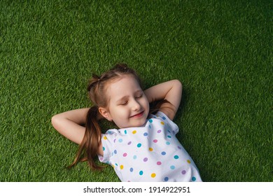 Cute Little Girl Lying On The Green Carpet, Lawn. The Child Is Resting, Lying On The Grass With His Eyes Closed. The Concept Of A Carefree Childhood, A Long-awaited Summer.