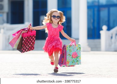 Cute Little Girl On Shopping. Portrait Of A Kid With Shopping Bags. Child In Dress, Sunglasses And Shoes Near Shopping Mall Having Fun. Shopping. Girl. 