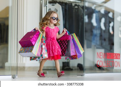 Cute Little Girl On Shopping. Portrait Of A Kid With Shopping Bags. Child In Dress, Sunglasses And Shoes Near Shopping Mall Having Fun. Shopping. Girl. 