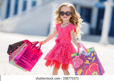 Cute Little Girl On Shopping. Portrait Of A Kid With Shopping Bags. Child In Dress, Sunglasses And Shoes Near Shopping Mall Having Fun. Shopping. Girl. 