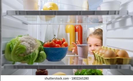 Cute Little Girl Opens Fridge Door, Looks Inside Takes Out Healthy Yogurt. Smart Little Child Eats Healthy. Shot From Inside The Fridge. Point Of View POV Shot From Refrigerator Full Of Good Food