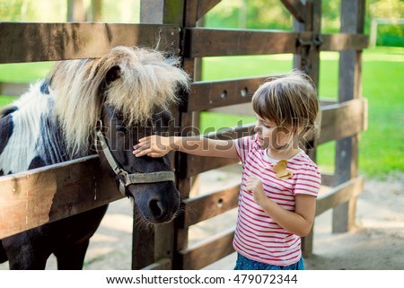 Cute little girl petting a pony in the zoo through a wooden fence