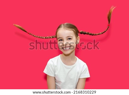 A cute little girl with pigtails on a red background. Portrait of a funny girl who waves her pigtails and laughs. Carefree childhood of a playful girl. The child has fun and throws pigtails