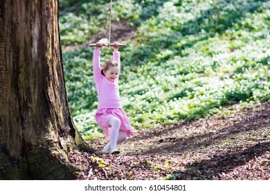 Cute Little Girl With Pink Dress On Tree Rope Swing In Blooming Spring Garden With Flowers. Child Swinging Outdoors In Backyard Playground. Kids Play Outdoor In Summer. Children Playing In Sunny Park
