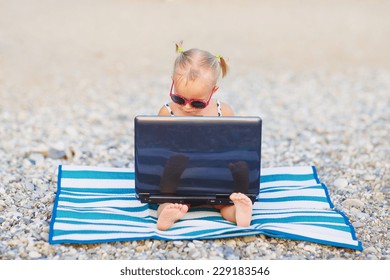 A Cute Little Girl In Pink Sunglasses Sitting On The Striped Mat With A Laptop On The Beach On A Warm Summer Day. Holiday On A Seashore Concept. Funny Kids.