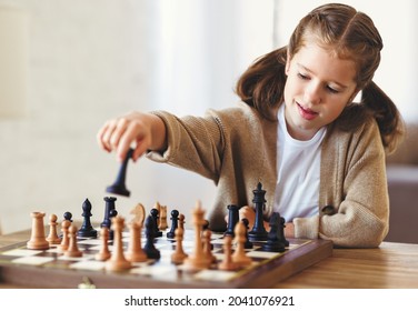 Cute little girl playing chess while spending leisure time at home during during day time, child sitting at table in living room and moving figure on chessboard with concentrated face expression