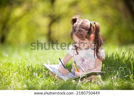 Cute little girl playing drawing in a sunny park. Beauty nature scene with colorful background at summer season. Family outdoor lifestyle. Happy girl relax on green grass