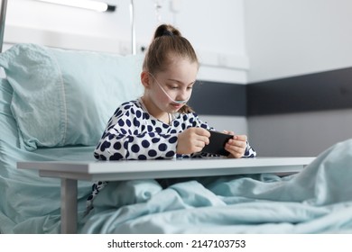 Cute Little Girl Playing Games On Smartphone While Resting On Bed In Pediatric Hospital Room. Joyful Young Patient Enjoying Mobile Phone Entertainment While Sitting In Clinic Recovery Ward.