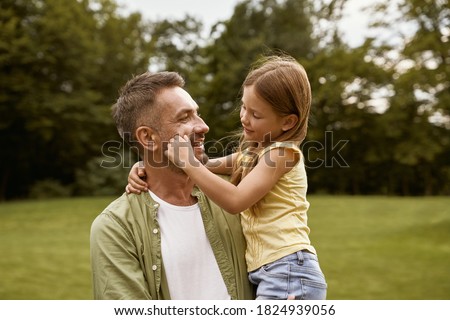Cute little girl playing with her loving father while visiting park on a summer day, touching his cheeks and smiling