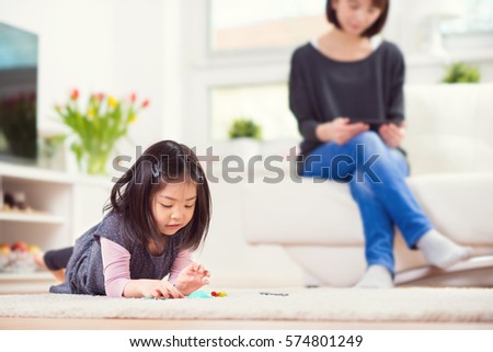 Cute little girl playing on carpet and mother sitting on sofa with tablet in background