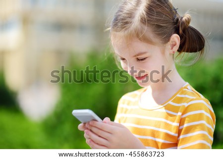 Cute little girl playing outdoor mobile game on her smart phone. Kid catching virtual pocket monsters. Modern addictive multiplayer location-based games.