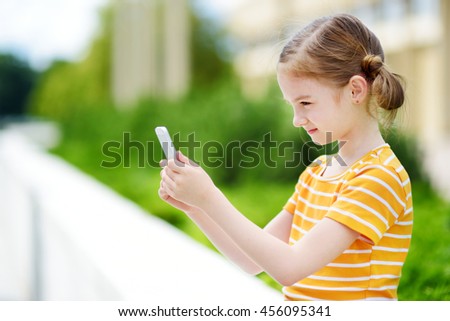 Cute little girl playing outdoor mobile game on her smart phone. Kid catching virtual pocket monsters. Modern addictive multiplayer location-based games.