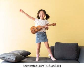 Cute Little Girl Playing Ukulele On Sofa With Fun, Inspiration For Kids Want To Be Musician Star