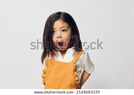 a cute little girl of preschool age is standing in an orange dress on a white background cheerfully contorting her face while standing sideways looking at the camera with her hands behind her back