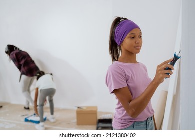 Cute Little Girl In Purple T-shirt And Hair Band Is Painting The Wall With A Roller That Is Covered With White Paint. Renovating A Room In The House. In The Background Mom And Sister Are Also Working