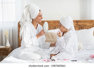 Cute Little Girl Putting Lipstick On Her Lips And Looking At Mirror In Her Mother Hands, Side View. Cheerful Beautiful Mom And Daughter Wearing Bathrobes, Having Beauty Day Together At Home