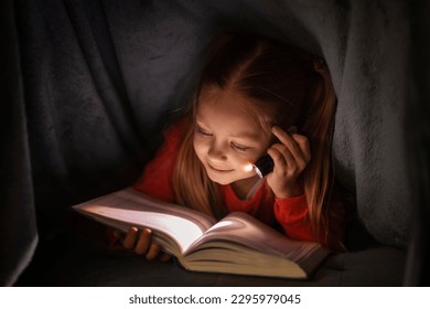 Cute little girl reading book while hiding under blanket with flashlight, happy preteen female child relaxing under covers, using torch in the dark, enjoying her favourite fairy tale, closeup