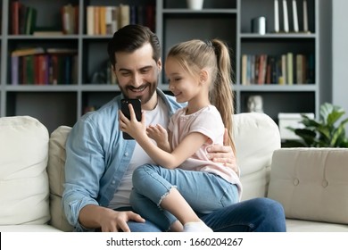 Cute little girl relax in living room with young dad show video on modern cellphone, happy father rest on sofa with small preschooler daughter using smartphone gadget together, technology concept - Powered by Shutterstock