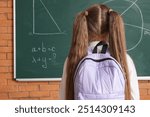 Cute little girl in school uniform with backpack near chalkboard in classroom, back view